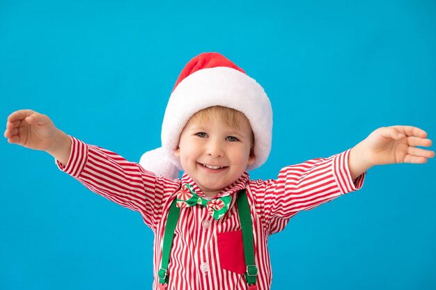 Lustiges Kind kleidete Weihnachtsmann-Hut gegen blauen Hintergrund an. Glückliches Kind, das Spaß zu Hause hat. Weihnachtsferienkonzept