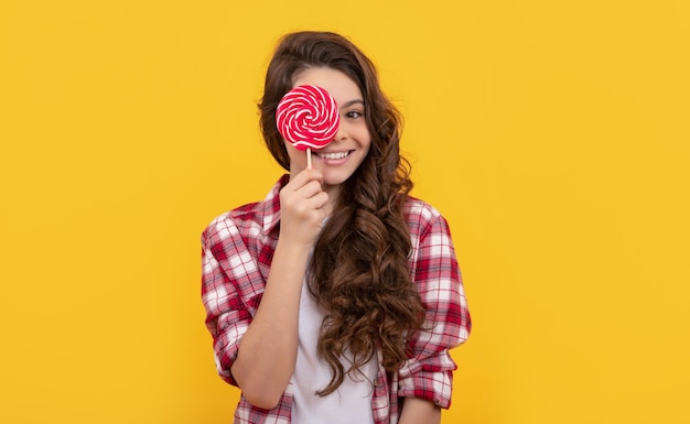 Lustiges Kind mit langen lockigen Haaren im Hemd hält Lollipop-Karamellbonbons auf gelbem Hintergrund lecker