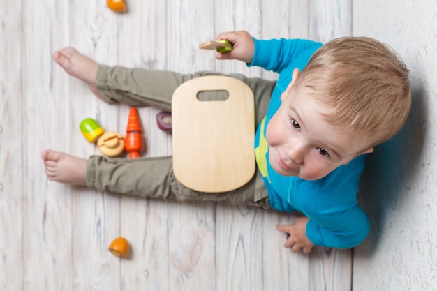 Lustiges Kind spielt im Chef. lächelndes Baby schneidet hölzernes Gemüse. Interessantes, sich sicher entwickelndes Kinderspiel hautnah.