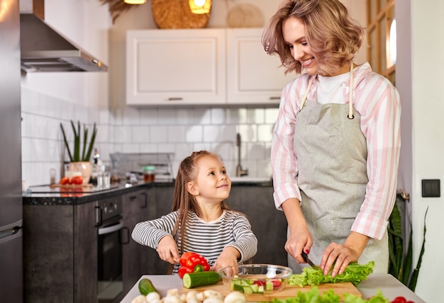 Lustiges Kindermädchen, das mit ihrer Mutter einen Salat in der modernen hellen Küche zubereitet, genießt den Prozess der Essenszubereitung