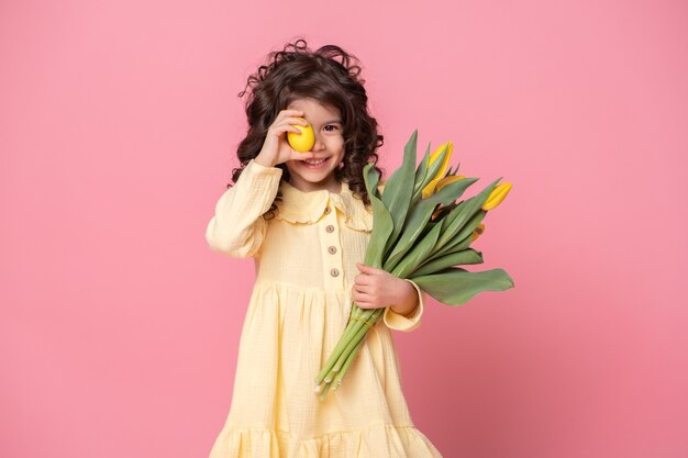 Lustiges Kindermädchen, das Tulpen und buntes Osterei vor ihrem Auge auf rosa Hintergrund hält.