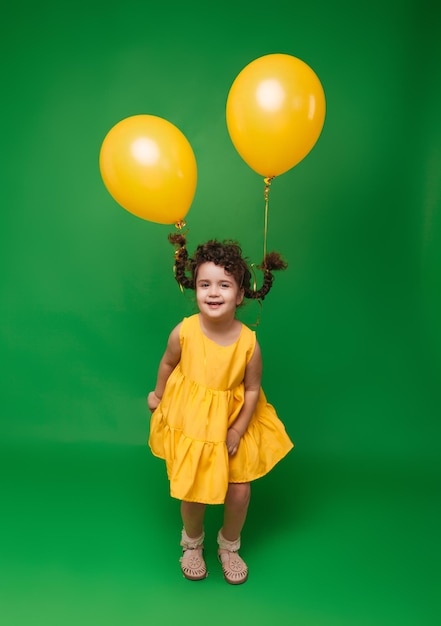 Lustiges kleines Mädchen auf grünem Hintergrund Ein Mädchen hat Spaß mit gelben Luftballons Luftballons heben die Zöpfe des Kindes an