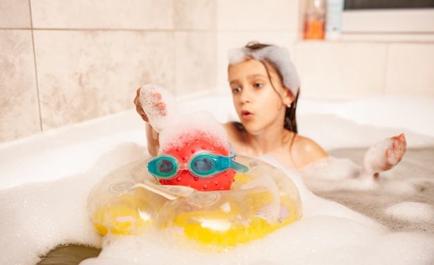 Lustiges kleines Mädchen badet in einer Badewanne mit Schaum und spielt lebensrettenden Ball