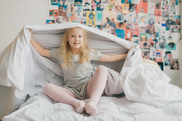 Lustiges kleines Mädchen, das auf weißem Bett sitzt und Decke über ihrem Kopf hält