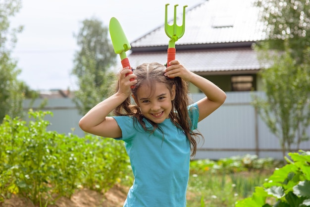 Lustiges kleines Mädchen, das kleine Gartengeräte hält, die im Garten stehen