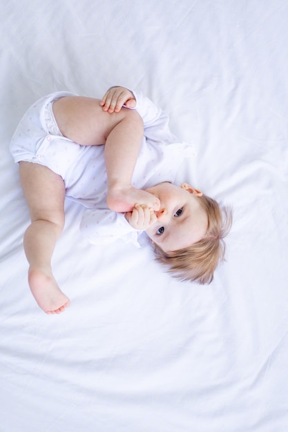 Lustiges kleines Mädchen, das mit ihren Füßen auf dem Bett in weißen Kleidern spielt und von oben lächelt, ein kleines Kind auf einem Baumwollbett zu Hause wachte morgens auf, nachdem es das Konzept der Kinderwaren geschlafen hatte