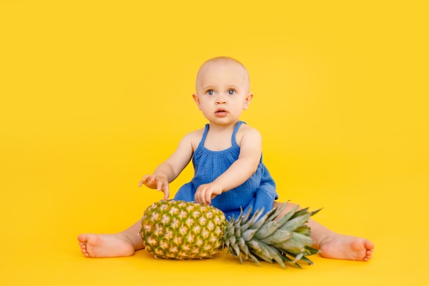 Lustiges kleines Mädchen gekleidet im blauen Kleid sitzend und spielend mit Ananas lokalisiert auf gelbem Hintergrund. Sommerferienkonzept