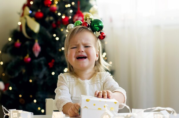 Lustiges kleines Mädchen im weißen Kleid, das zu Hause in der Nähe des Weihnachtsbaums Weihnachtsgeschenkbox öffnet