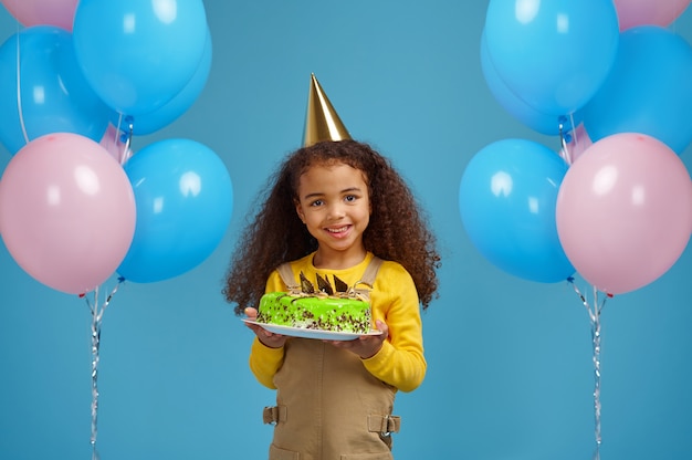 Lustiges kleines Mädchen in der Mütze hält Geburtstagskuchen, blauer Hintergrund. Hübsches Kind hat eine Überraschung, Eventfeier, Ballondekoration bekommen