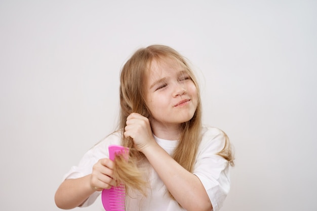 Lustiges kleines Mädchen kämmt langes und wirres Haar auf weißem Hintergrund. Kosmetika zur Pflege von Kinderhaaren.