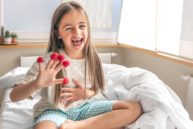 Lustiges kleines Mädchen mit Himbeeren an den Fingern morgens im Bett