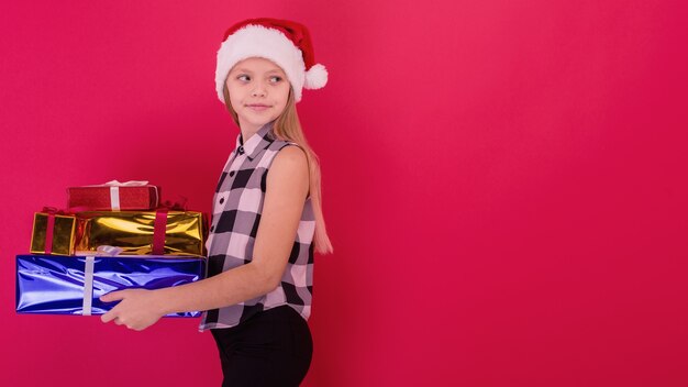 Lustiges lächelndes fröhliches Kindermädchen in Sankt-Hut, das Weihnachtsgeschenk in der Hand über dem roten Hintergrund hält