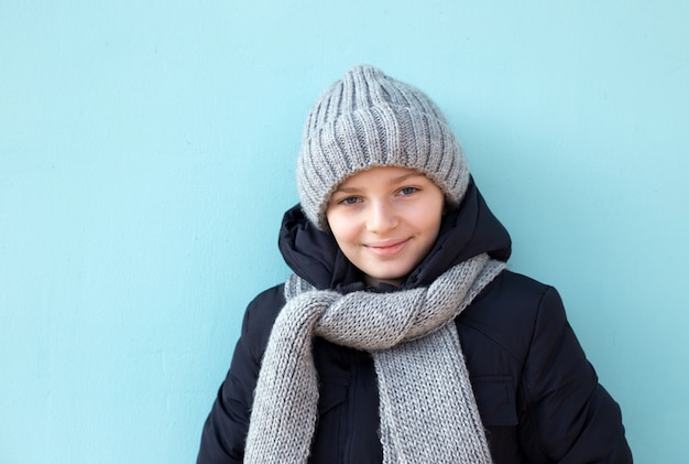 Lustiges lächelndes Kind bereit zu den Winterferien, zum modernen Jungen in der grauen Kappe des Winters und zum Schal, der gegen blaue Wand steht