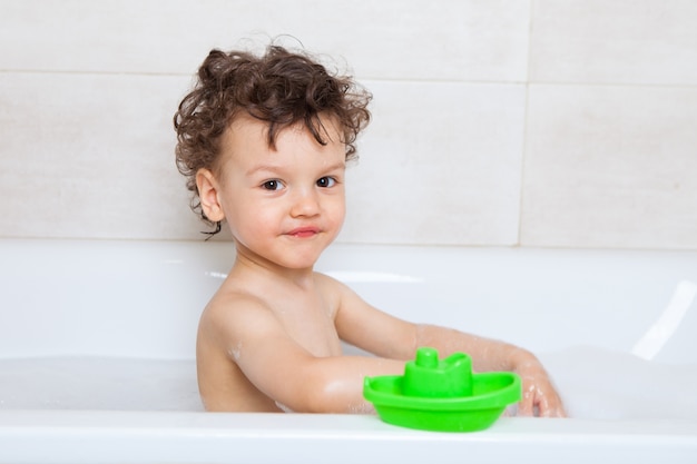 Lustiges lockiges Baby badet in einem Bad mit einem grünen Schiff. Persönliche Hygieneregeln für Kinder. Junge, ein Kind spielt mit Spielzeug in einem Vollbad mit Wasser.