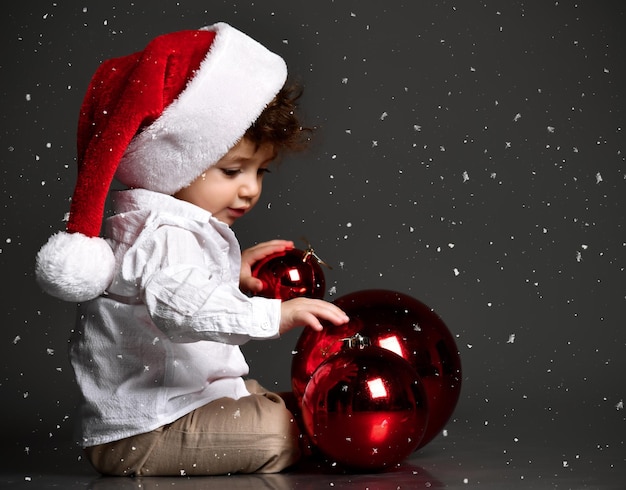 Lustiges lockiges Baby in Weihnachtsmütze, das mit weihnachtlichen roten Kugeln auf dunklem Studiohintergrund spielt Neujahrskarte erster Weihnachtsschnee