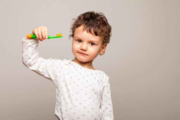 Lustiges lockiges Kind steht lächelnd und hält eine Zahnbürste in seinen Händen. Gesunde starke Kinderzähne. Die Regeln der persönlichen Hygiene.