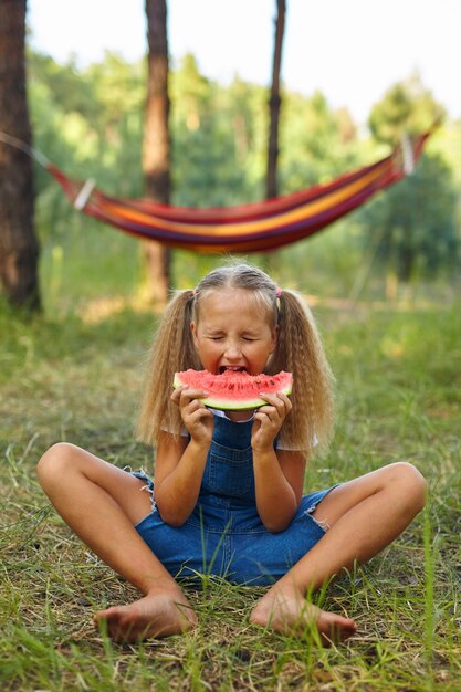 Lustiges Mädchen, das Wassermelone im Park isst. Kind isst Obst im Freien. Gesunder Snack für Kinder. Kleines Mädchen, das im Wald spielt und eine Scheibe Wassermelone beißt