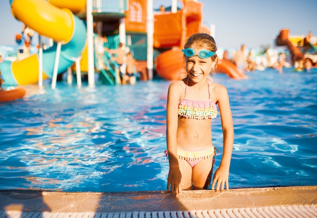 Lustiges Mädchen in der blauen Schutzbrille zum Schwimmen steht in einem Pool mit sauberem transparentem Wasser, das in die Kamera schaut und die warme Sommersonne genießt