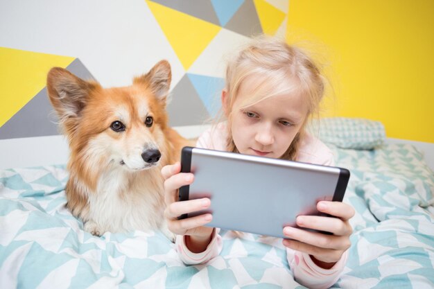 Lustiges Mädchen und Hundecorgi auf dem Bett im Kinderzimmer lasen ein Buch