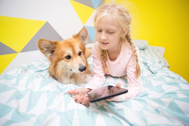 Lustiges Mädchen und Hundecorgi auf dem Bett im Kinderzimmer lasen ein Buch