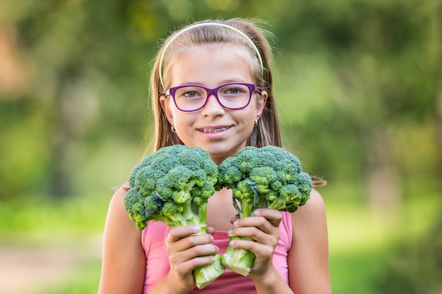 Lustiges nettes Mädchen, das in den Händen Rotkohl und Brokkoli hält. Unscharfer Hintergrund im Garten. Pre-Teen junges Mädchen mit Brille und Zahnspangen.
