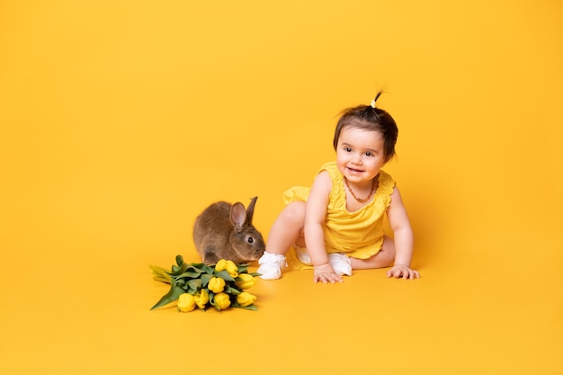 Lustiges niedliches Baby im gelben Kleid mit Kaninchen und Tulpen, die auf gelbem Hintergrund sitzen.