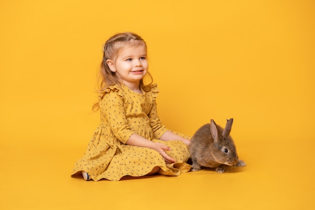 Lustiges niedliches Kindermädchen im gelben Kleid mit Kaninchen, das auf gelbem Hintergrund sitzt.