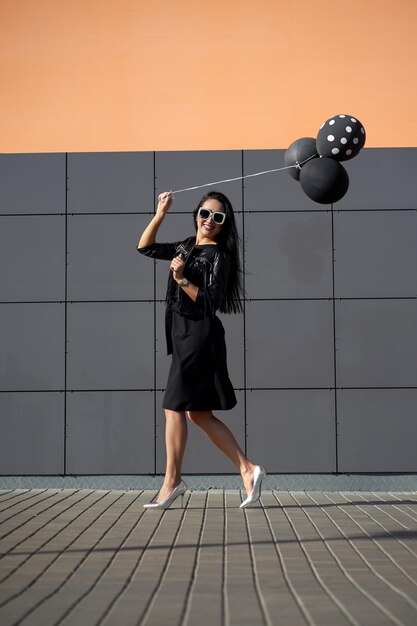 Lustiges positives attraktives Modell mit charmantem Lächeln in stilvoller Sonnenbrille, die elegantes Kleid trägt, modische schwarze Lederjacke mit Fransen, die schwarze Ballons die Hand hält. Model im trendigen Outfit.