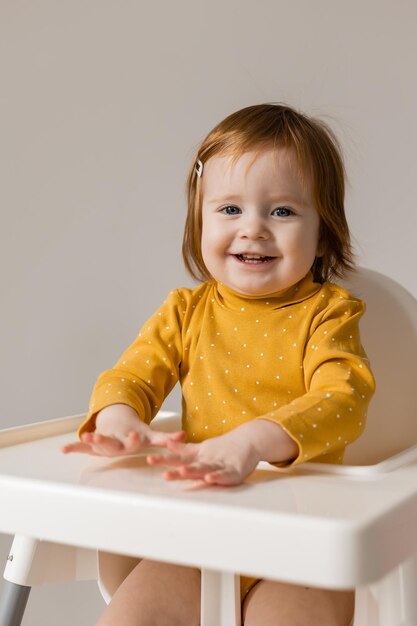 Lustiges rothaariges blauäugiges Baby in einem gelben Body, der in einem weißen Hochstuhl sitzt