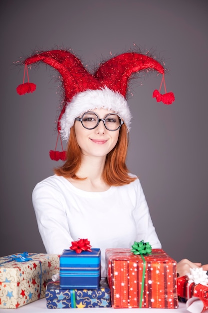 Lustiges rothaariges Mädchen in der Weihnachtsmütze und in den Gläsern mit Geschenkboxen.