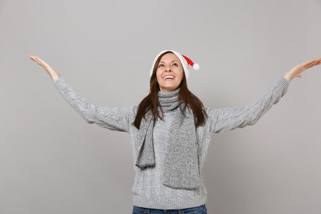 Lustiges Sankt-Mädchen in grauem Pullover-Schal-Weihnachtshut, der nach oben schaut und zeigende Hände beiseite lokalisiert auf grauem Hintergrund ausbreitet. Frohes neues Jahr 2019 Feier Urlaub Party Konzept. Kopieren Sie Platz.