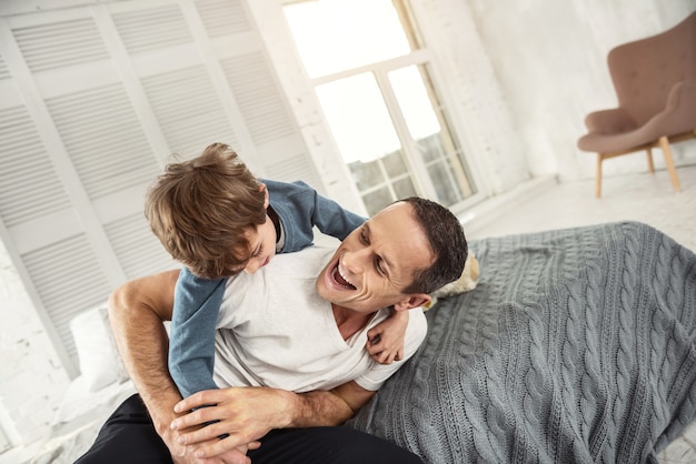 Lustiges Spiel. Attraktiver fröhlicher dunkelhaariger Papa, der mit seinem Sohn lacht und auf dem Bett liegt und Spaß hat