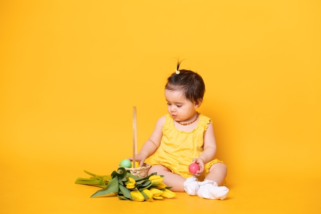 Lustiges süßes Baby im gelben Kleid mit Tulpen, Korb mit bunten Eiern.