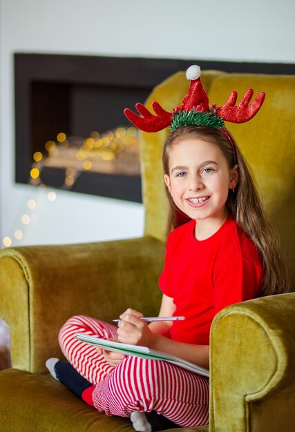 Lustiges süßes kaukasisches Mädchen mit Linkshänder schreibt einen Brief an den Weihnachtsmann in der Nähe des Weihnachtsbaums