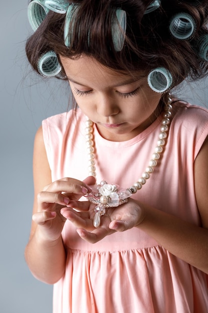 Lustiges süßes kleines Kindermädchen in rosa Kleid und Lockenwicklern schaut auf die Halskette