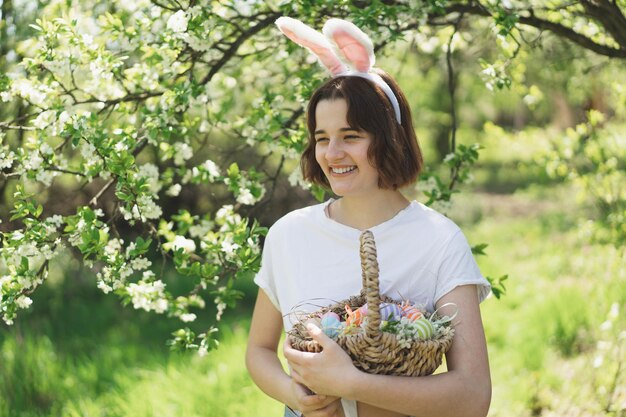 Lustiges Teenager-Mädchen mit Eierkorb und Hasenohren auf Ostereiersuche im sonnigen Frühlingsgarten