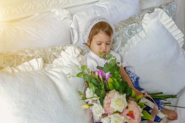 Lustiges und süßes brünettes kleines lächelndes Mädchen, das mit Blumenstrauß spielt, der sich auf dem Bett im hellweißen Schlafzimmer zu Hause entspannt. Weißes Interieur mit großem Bett. Kindheit, Vorschule, Jugend, Entspannungskonzept
