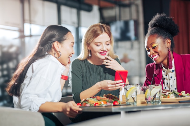 Lustiges Video ansehen Drei Geschäftsfrauen, die lustiges Video auf rotem Smartphone ansehen, das im Restaurant sitzt