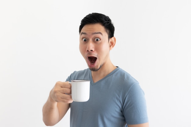Lustiges Wowgesicht des asiatischen Mannes im blauen T-Shirt trinkt Kaffee vom weißen Becher.