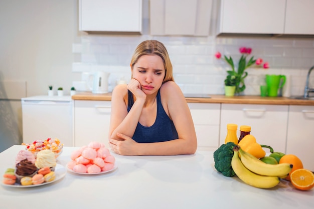 Luta com dieta. Jovem mulher triste em t-shirt azul, escolhendo entre legumes frescos ou doces na cozinha. Escolha entre alimentos saudáveis e não saudáveis. Dieta. Comida saudável