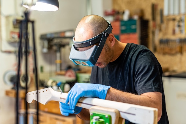 Luthier en el trabajo con lentes de precisión, artesano experto en la fabricación de instrumentos musicales con mango de guitarra, concepto de negocio musical y artesanal, uso de protección en el lugar de trabajo