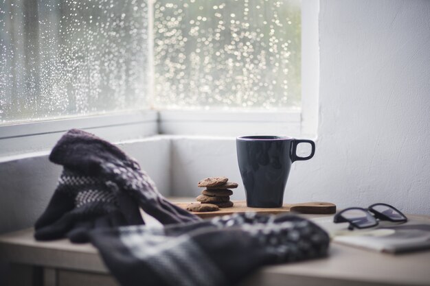 Luvas de inverno e gorro com uma xícara de café e biscoito na mesa de madeira para o fundo do inverno