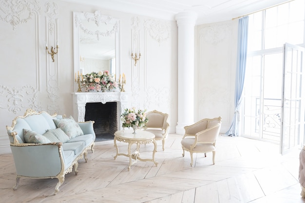 Luxuoso interior claro em estilo barroco. Um quarto espaçoso com uma bela mobília elegante, lareira e flores. estuque de plantas nas paredes e parquet de madeira clara