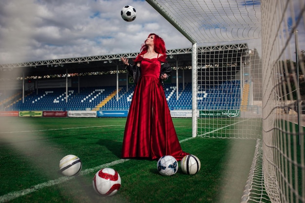 Luxuriöse Frau mit roten Haaren und in einem roten Kleid spielt auf dem Fußballplatz Idee und Konzept einer Kombination aus Sport und Schönheit ungewöhnlicher Präsentation