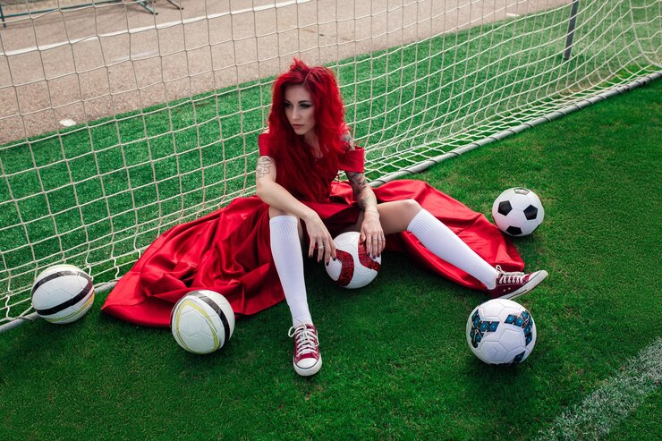 Luxuriöse Frau mit roten Haaren und in einem roten Kleid spielt auf dem Fußballplatz Idee und Konzept einer Kombination aus Sport und Schönheit ungewöhnlicher Präsentation