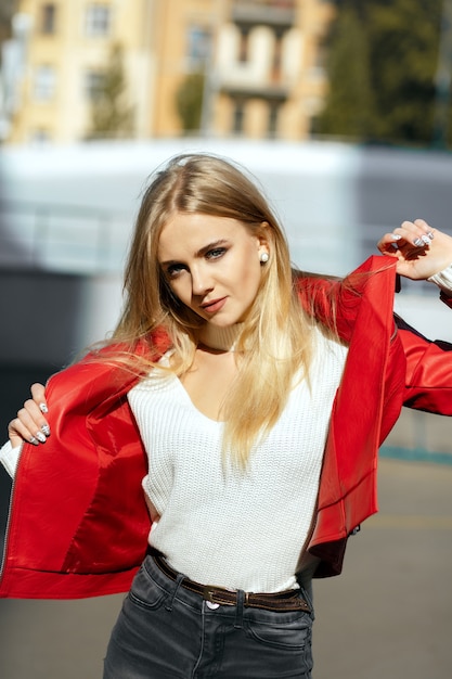 Luxuriöses blondes Model mit langen Haaren in roter Lederjacke posiert im Sonnenlicht