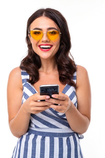 Luxuriöses brünettes Mädchen mit schönem Make-up in einem gestreiften Kleid und Brille mit einem Telefon in ihren Händen auf einem weißen Hintergrund.