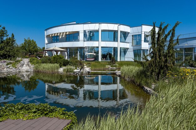 Luxuriöses, stilvolles, weißes, zweistöckiges Landhaus mit abgerundeten Formen und breiten Fenstern, die sich an einem sonnigen Sommertag im Wasser eines kleinen künstlichen Teiches im Hinterhof widerspiegeln