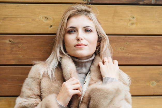 Luxus blonde Frau mit blauen Augen und langen Haaren im Pelzmantel 30 Jahre alt auf Holzwand.