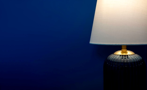 Luxus-Tischlampe in Nahaufnahme mit weißem Stoff, blauem Glas und goldfarbenen Materialien, die auf dunkelblauem Wandhintergrund mit Kopierraum leuchten Beleuchtungslampe im Dunkeln